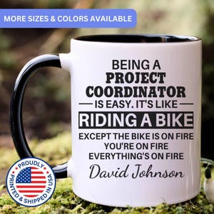 May include: A white ceramic mug with a black interior and handle, featuring black text that reads: BEING A PROJECT COORDINATOR IS EASY. IT'S LIKE RIDING A BIKE EXCEPT THE BIKE IS ON FIRE YOU'RE ON FIRE EVERYTHING'S ON FIRE David Johnson.