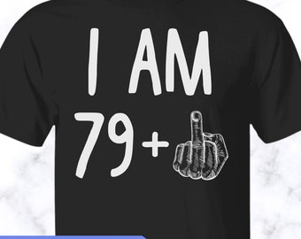 May include: A black short-sleeved t-shirt featuring the white text I AM 79+ and a white graphic illustration of a hand making a middle finger gesture. The shirt includes a blue rectangular banner at the bottom with the text MORE STYLES & COLORS AVAILABLE!