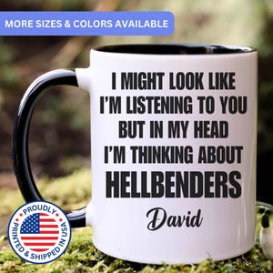 May include: A white ceramic mug with a black interior and handle. The mug features black text that reads: I MIGHT LOOK LIKE I'M LISTENING TO YOU BUT IN MY HEAD I'M THINKING ABOUT HELLBENDERS, followed by the name David in a script font.