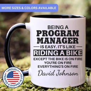 May include: White ceramic mug with a black handle and interior. The mug features the text "BEING A PROGRAM MANAGER IS EASY. IT'S LIKE RIDING A BIKE EXCEPT THE BIKE IS ON FIRE YOU'RE ON FIRE EVERYTHING'S ON FIRE David Johnson".