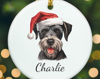 May include: A round white ceramic ornament featuring a detailed illustration of a black and gray Schnauzer dog wearing a red and white Santa hat. The dog has its mouth open with its tongue visible. The name Charlie is printed in black script below.