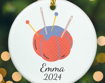 May include: A round white ceramic ornament featuring a graphic illustration of a coral-colored pincushion with a blue top, accented by several sewing needles and pins. The name Emma and the year 2024 are printed in black text below the illustration.