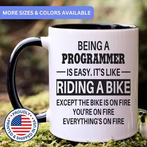 May include: A white ceramic mug with a black handle and interior, featuring black text that reads: BEING A PROGRAMMER IS EASY. IT'S LIKE RIDING A BIKE EXCEPT THE BIKE IS ON FIRE YOU'RE ON FIRE EVERYTHING'S ON FIRE. A circular logo with a flag is present.