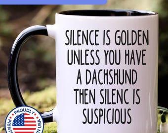 May include: A white ceramic mug with a black handle and interior, featuring the black printed text: SILENCE IS GOLDEN UNLESS YOU HAVE A DACHSHUND THEN SILENC IS SUSPICIOUS. A circular badge with the American flag and text is visible on the lower left.