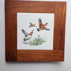 May include: A framed ceramic tile with a painted image of three brown and white quail birds in flight against a white background. The frame is made of dark brown wood.