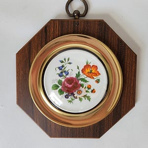 May include: A vintage wall hanging featuring a floral design. The centerpiece is a white ceramic plate with a colorful arrangement of flowers, framed by a brass ring and a dark wood octagonal frame. A brass ring is at the top for hanging.