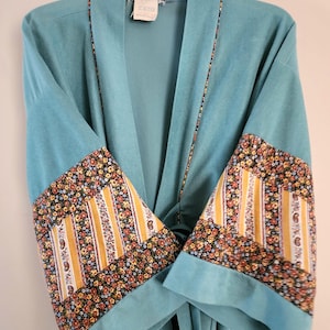 May include: A turquoise robe with floral patterned sleeves and trim. The sleeves feature a design of yellow stripes and small flowers. The robe is hanging on a white hanger.