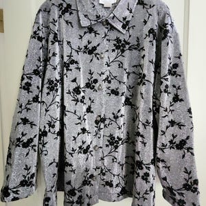 May include: A silver button-down shirt with a black floral pattern. The long-sleeved shirt has a collared neckline and is hanging on a black hanger. The fabric has a subtle shimmer.