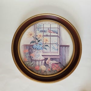 May include: Round framed print featuring a still life scene. The artwork depicts a window view with flowers, fruit, and a pitcher. The frame is brown with a gold inner edge. The print has a vintage aesthetic.