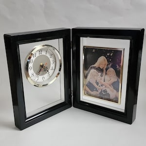May include: A hinged, dual-panel black frame featuring a circular analog clock with a white face and black numerals on the left side, and a rectangular photo frame containing a picture of three people on the right side.
