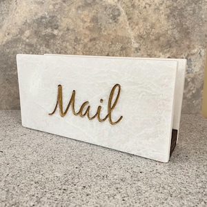 May include: A rectangular mail holder featuring a white marble-patterned exterior with a smooth finish. The front panel displays the word Mail in a raised, metallic gold cursive script font. The structure is open at the top and sides for storage.