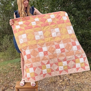 May include: A square quilt featuring a geometric patchwork pattern in shades of peach, coral, yellow, and white. The design consists of repeating star and block motifs arranged in a grid, creating a soft, warm colour gradient across the fabric surface.