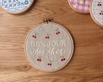 May include: A circular wooden embroidery hoop containing beige fabric with white embroidered text that reads BONJOUR Mon chéri, surrounded by small red cherry motifs and white French knots. Other hoops with floral, bear, and gingham patterns are visible.