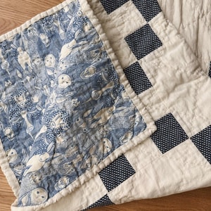 May include: A quilted blanket featuring a blue and white fabric with a repeating pattern of various woodland animals including owls and rabbits, contrasted with a cream and dark blue checkered border, showcasing detailed stitching throughout.