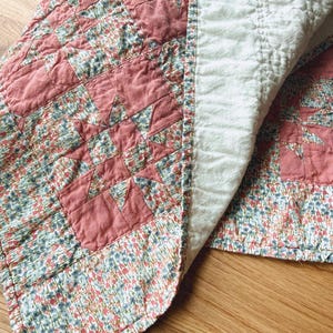 Könnte beinhalten: Eine Steppdecke mit einem Patchwork-Design aus einfarbigen, altrosa Stoffquadraten und floral gemusterten Stoffquadraten mit blauen, rosa und grünen Blumen. Die Rückseite besteht aus einfarbigem, cremeweißem Stoff mit sichtbaren gitterartigen Nähten.