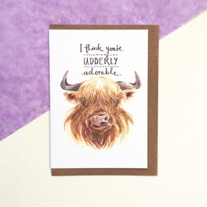 May include: A greeting card with a watercolor illustration of a Highland cow. The card features the text "I think you're udderly adorable" in black script. The card has a brown border and is set against a purple and white background.