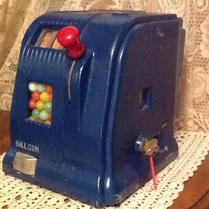 May include: A blue vintage ball gun toy with a red handle and a clear plastic container filled with colorful balls. The toy has a label that reads "BALL GUN".