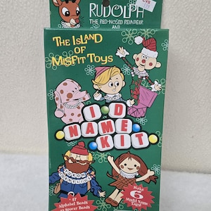 May include: A green box featuring illustrations of characters from "Rudolph the Red-Nosed Reindeer" and "The Island of Misfit Toys." The box is labeled "I.D. Name Kit" and includes alphabet beads, spacer beads, a bead tray, and elastic string to make six misfit name tags.