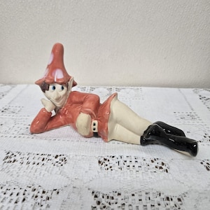 May include: A ceramic figurine of a reclining elf. The elf wears a red jacket, a white skirt, and black boots. The elf has a red hat with white spots and is resting their head on their hand.