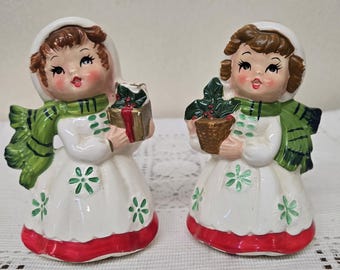 Op de afbeelding: Twee keramische beeldjes van meisjes in witte jurken met groene sjaals en rode accenten. Eén houdt een cadeau vast, de ander een potplant. De beeldjes hebben bruin haar en dragen witte mutsen. De jurken hebben groene bloemdetails.