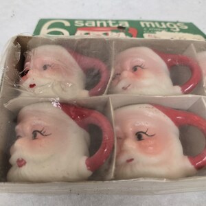 May include: A set of six Santa mugs in a cardboard box. The mugs are shaped like Santa's face, with white beards, rosy cheeks, and red hats and handles. The box has the words "6 santa mugs" printed on it.
