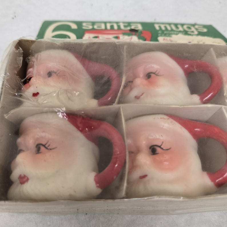 May include: A set of six Santa mugs in a cardboard box. The mugs are shaped like Santa's face, with white beards, rosy cheeks, and red hats and handles. The box has the words "6 santa mugs" printed on it.