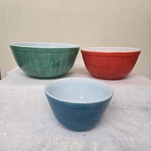 May include: Three vintage mixing bowls in shades of green, red, and blue. The bowls have a classic rounded shape with white interiors. The green bowl has a marbled effect. A set of kitchenware.