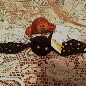May include: A plush black bat toy with wide wings featuring colorful polka dots and a small white tag attached with a yellow ribbon, positioned in front of a smaller, round, textured copper-colored plush creature with large dark eyes.