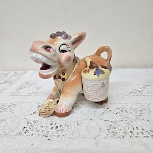 May include: A ceramic salt shaker shaped like a brown and white donkey carrying a basket labeled "S". The donkey has a purple flower on its head and a yellow rope around its neck.