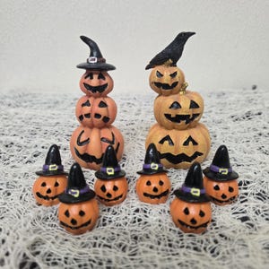 May include: A collection of Halloween-themed decorations. The image features several orange pumpkin figurines with carved faces. Some pumpkins are stacked, and others wear witch hats. A black crow sits atop one pumpkin stack.
