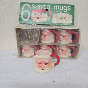 May include: A set of six Santa mugs in a box. The mugs are white with red handles and Santa faces. The box is green and white and reads "6 santa mugs". The mugs are designed to be used as candle holders, for cigarettes, candy, and party use.