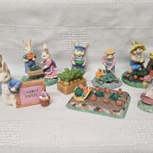 May include: A collection of Easter bunny figurines in various poses, including gardening and carrying vegetables. The figurines are painted in pastel colors and feature detailed clothing and accessories. One figurine holds a sign that reads "Carrot Patch".
