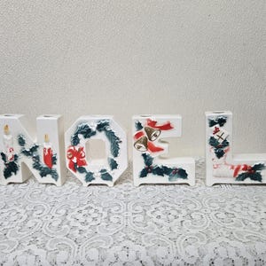 May include: White ceramic "NOEL" letters, each decorated with Christmas motifs. The "N" and "L" feature candles and holly, the "O" has a wreath, and the "E" displays bells and ribbons. Festive holiday decor.