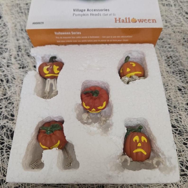 Peut inclure: Un ensemble de cinq figurines miniatures en forme de t&ecirc;tes de citrouille avec des jambes et des bras squelettiques, emball&eacute;es dans un plateau en polystyr&egrave;ne blanc. La bo&icirc;te indique Village Accessories Pumpkin Heads Set of 5 Halloween Series.