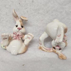 May include: Two ceramic Easter bunny figurines. One bunny is lying on its back with its paws up and a pink bow tie. The other bunny is upside down, with its head on the ground. Both bunnies are white with brown and pink accents.