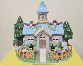 May include: A small, blue and white ceramic church figurine with a pink roof and a bell on the steeple. The church is surrounded by a white picket fence and green bushes with pink and yellow flowers. There are two small black lampposts on either side of the church.