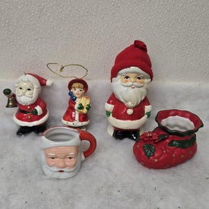 May include: A collection of Christmas-themed ceramic figurines. Includes a Santa Claus mug, a Santa Claus figure with a bell, a girl holding a gift, a Santa Claus figure, and a red boot-shaped planter.