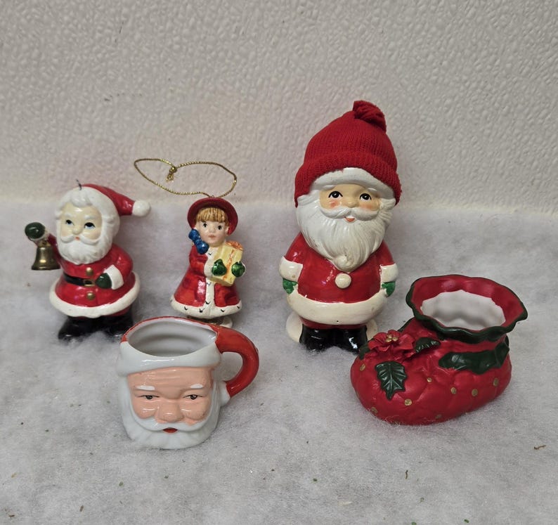 May include: A collection of Christmas-themed ceramic figurines. Includes a Santa Claus mug, a Santa Claus figure with a bell, a girl holding a gift, a Santa Claus figure, and a red boot-shaped planter.