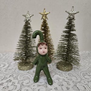 May include: A ceramic Christmas figurine of a child in a green jumpsuit with a curved hat. The figurine has a painted face with brown hair. Behind the figurine are three small Christmas trees with silver and gold star toppers.