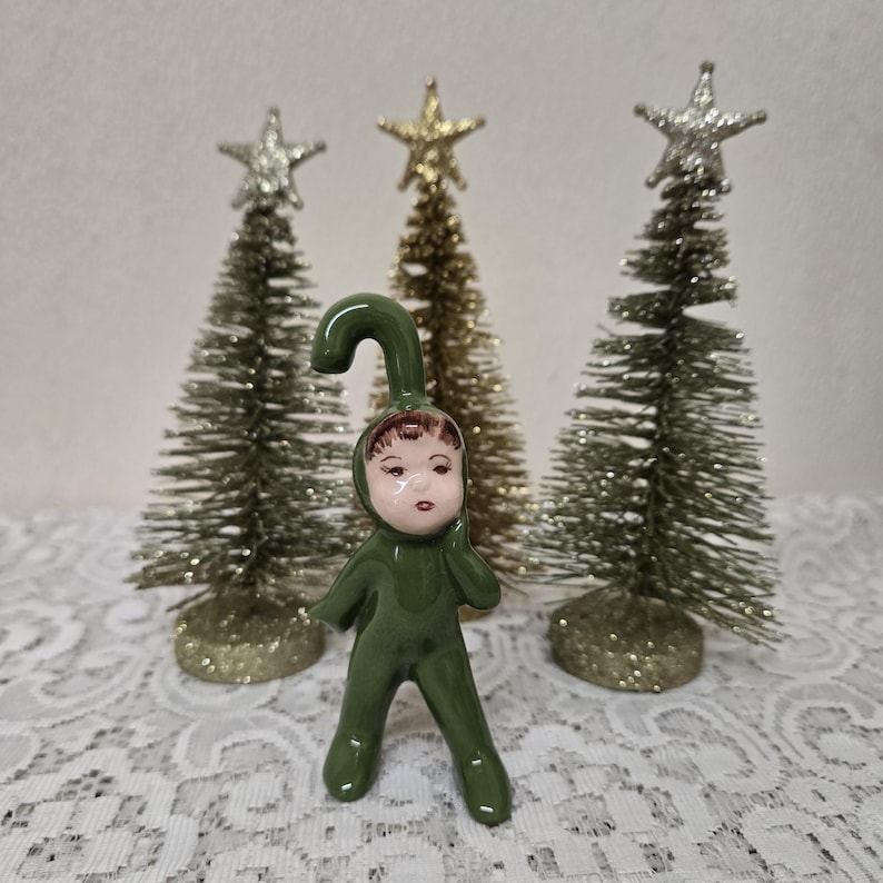 May include: A ceramic Christmas figurine of a child in a green jumpsuit with a curved hat. The figurine has a painted face with brown hair. Behind the figurine are three small Christmas trees with silver and gold star toppers.