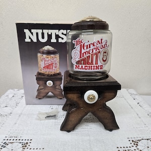 May include: A vintage glass jar with a dark wooden lid and base, labeled "The Great American Nut Machine." The jar sits on a small, decorative wooden stand with a white and gold knob. A box with the word "NUTS" is in the background.