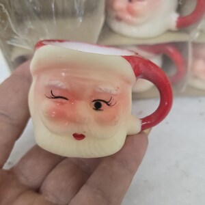 May include: A small, ceramic Santa Claus mug with a red handle and a winking face. The mug has a cream-colored base, a red rim, and a white beard. The mug is part of a set, with others visible in the background.