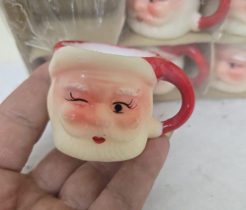 May include: A small, ceramic Santa Claus mug with a red handle and a winking face. The mug has a cream-colored base, a red rim, and a white beard. The mug is part of a set, with others visible in the background.