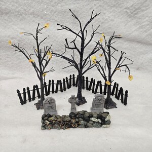 Peut inclure: Une sc&egrave;ne de cimeti&egrave;re miniature comprenant trois arbres noirs scintillants sans feuilles avec de petites feuilles jaunes, une cl&ocirc;ture noire, un mur en pierre et trois pierres tombales grises. Une pierre porte l'inscription Harry Wolfman 1889-1956.