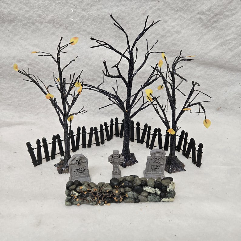 Peut inclure: Une sc&egrave;ne de cimeti&egrave;re miniature comprenant trois arbres noirs scintillants sans feuilles avec de petites feuilles jaunes, une cl&ocirc;ture noire, un mur en pierre et trois pierres tombales grises. Une pierre porte l'inscription Harry Wolfman 1889-1956.