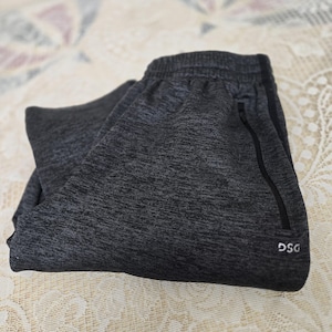 May include: Folded dark gray athletic pants with a drawstring waistband and a black zipper pocket. The pants have the letters "DSG" printed on the lower leg. The fabric has a heathered texture.