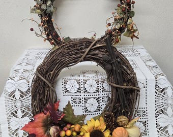 May include: A fall-themed wreath made of woven brown twigs, adorned with colorful autumn leaves, sunflowers, berries, and small pumpkins. The wreath is decorated with a sunflower-patterned bow and a pinecone. The wreath is displayed on a white lace tablecloth.
