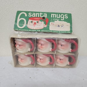 May include: A package of six Santa mugs. Each mug is white with a red handle and Santa's face. The package has a green label that reads "6 santa mugs" and "USE AS CANDLE HOLDERS, FOR CIGARETTES, CANDY & PARTY USE."