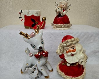 May include: A collection of vintage Christmas figurines, including a red boot, an angel, a reindeer with a red bow, and a winking Santa Claus. The figurines are white, red, and gold, with intricate details.