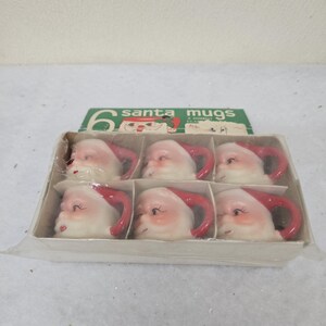 May include: A set of six Santa mugs in a cardboard box. Each mug is shaped like Santa Claus's face, with a red hat and handle. The mugs are white with pink cheeks and a smiling expression. The box has the text "santa mugs".